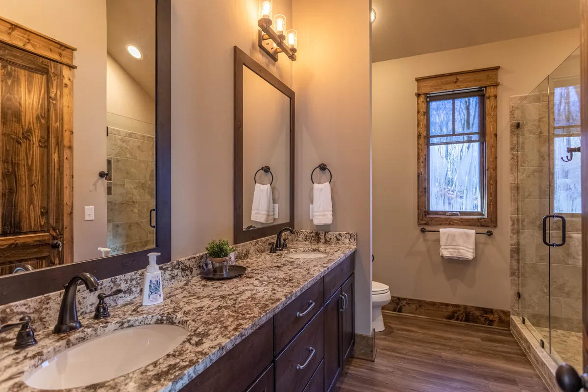 An elegant ensuite bathroom featuring a double granite vanity, large mirror, wooden door, and a glimpse of a luxurious walk-in shower. Early morning light streams through the window, elevating the spa-like experience of the space.