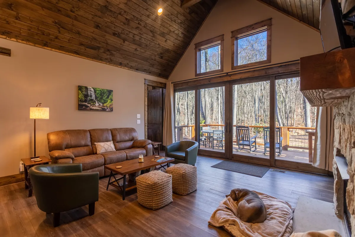 A warm and inviting living room with a comfortable leather sofa, a wooden coffee table, and a wall of glass doors that open to the outdoors. The room is filled with natural light, and the large windows offer glimpses of mountain scenery, creating a seamless connection between the interior and the surrounding nature.