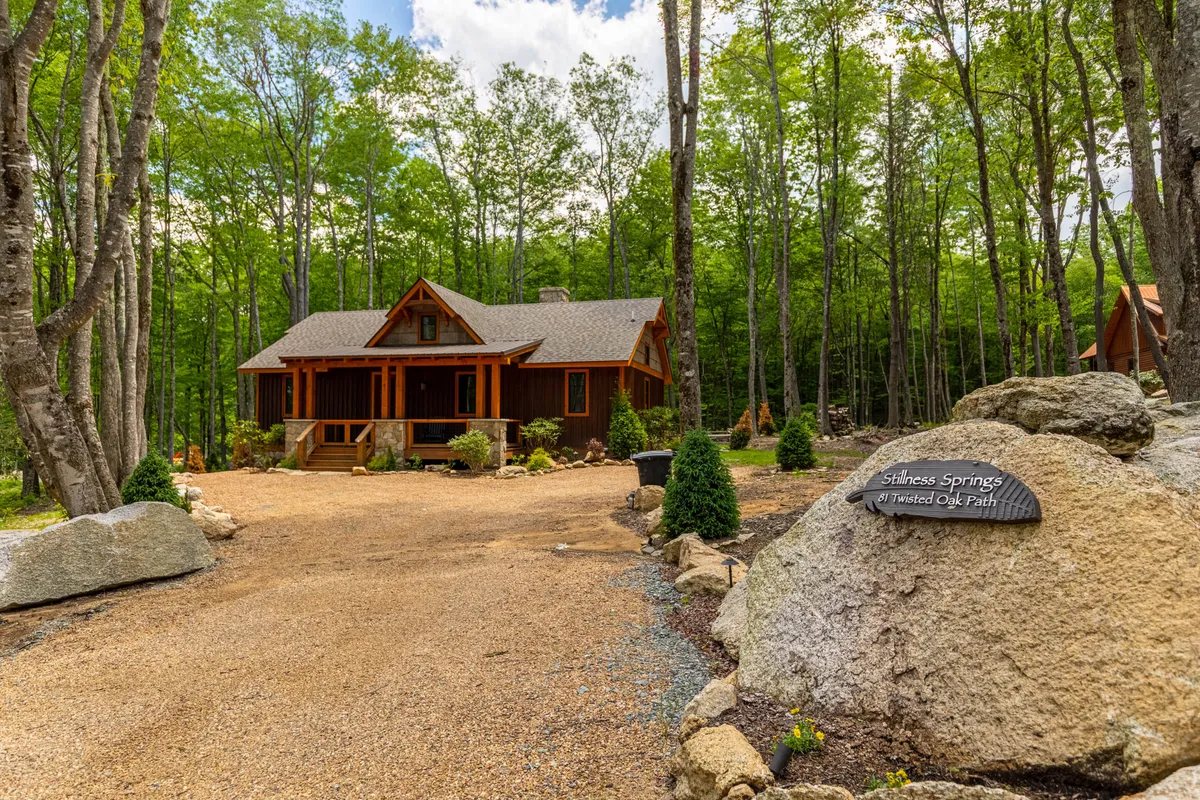 This exterior shot features a charming wooden cabin surrounded by mature landscaping, a rock sign at the entrance, and a wide driveway. The spacious grounds hint at ample room for outdoor activities and gatherings, with plenty of green space ideal for pets and children to play.