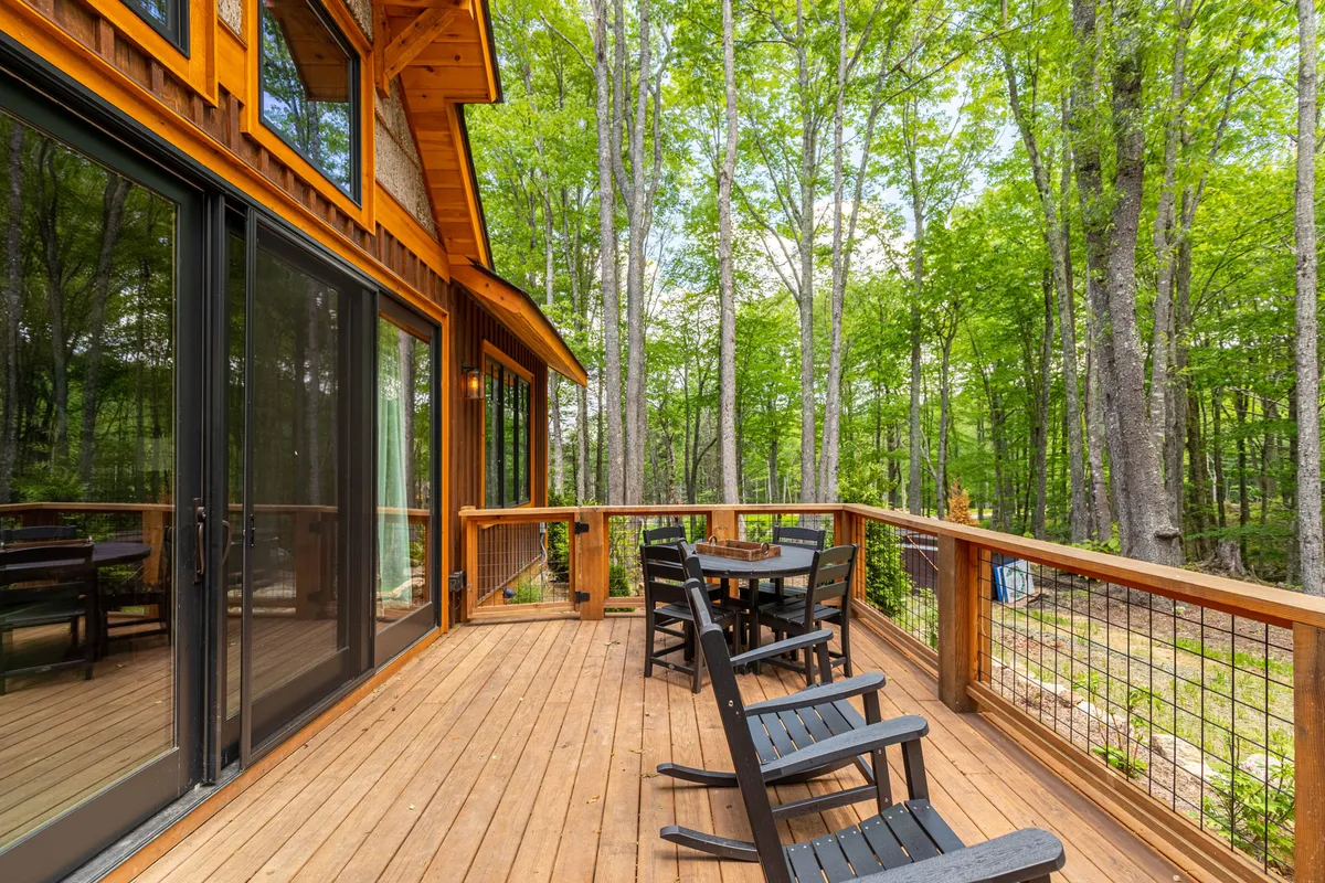 An inviting wooden deck with comfortable chairs sits nestled among tall trees, providing a tranquil spot to relax, savor coffee, and watch wildlife. The deck looks out onto the property's green surroundings, showcasing the natural setting.