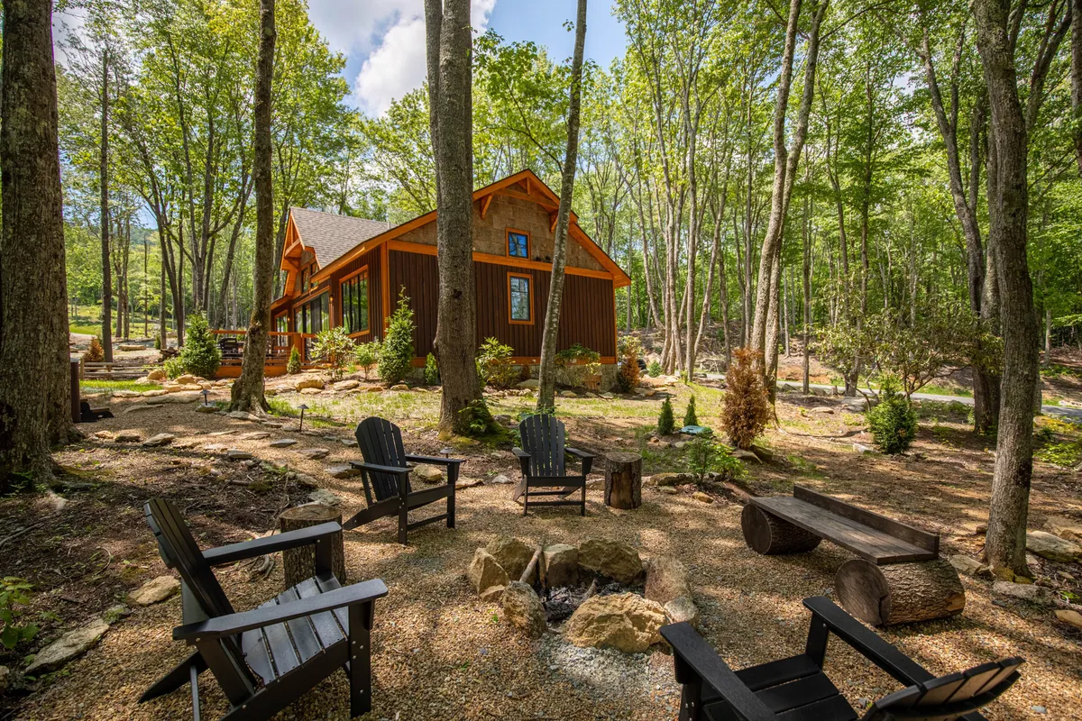 An inviting outdoor fire pit area with wooden chairs arranged for conversation. The fire pit is set among trees, with the house's facade visible in the background, evoking cozy evenings spent outdoors, perhaps with a glass of wine and good company.