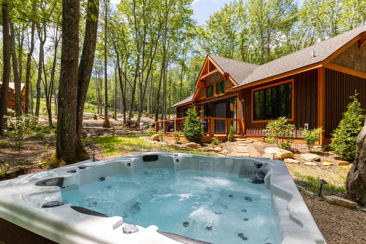 This image features a bubbling hot tub nestled next to a wooden cabin, surrounded by lush trees and foliage. The setting suggests privacy and immersion in nature, with the hot tub as the main focal point, inviting guests to relax outdoors while soaking in the tranquil mountain atmosphere.