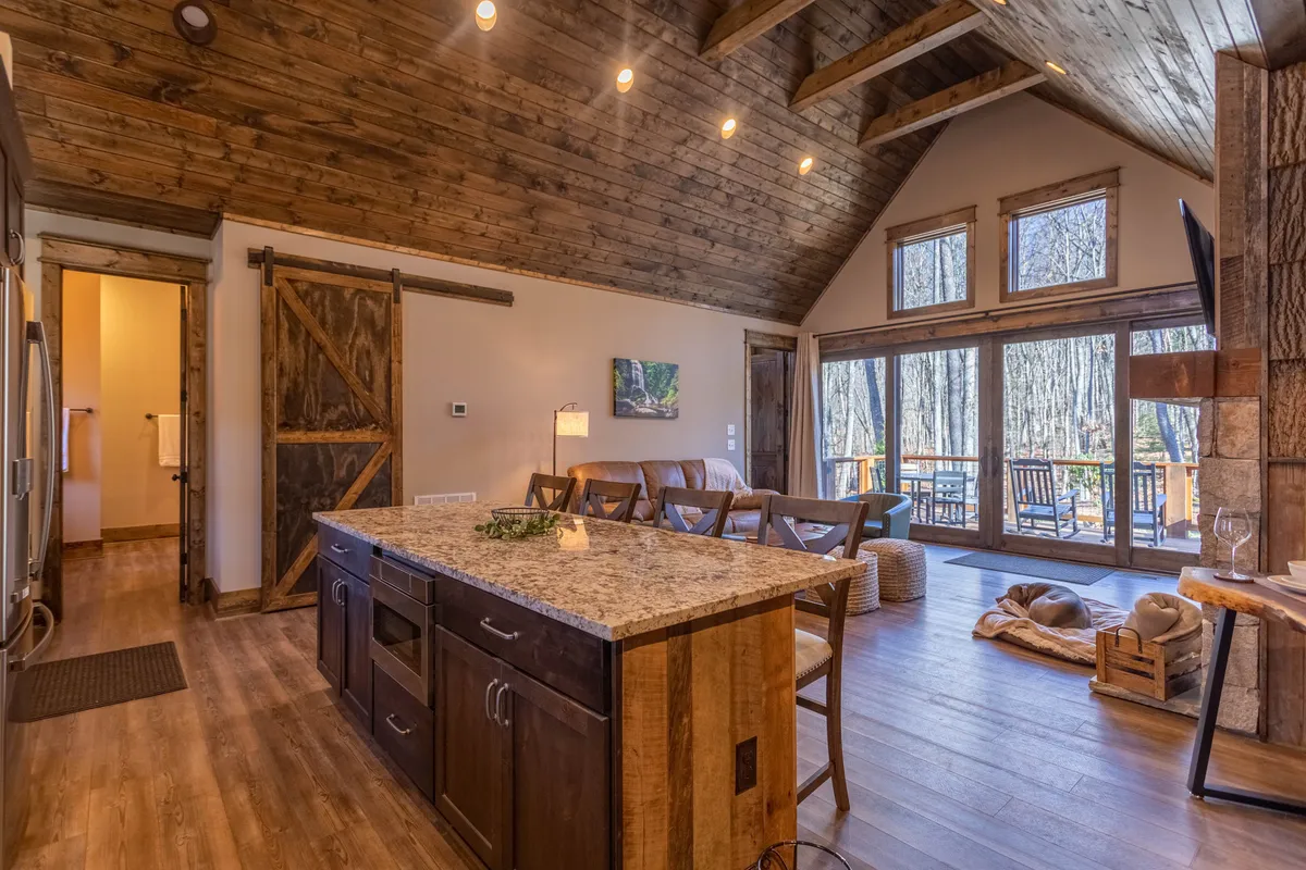 A spacious kitchen and lounge area featuring a kitchen island, wooden beams, large windows framing natural views, and comfortable seating. The warm timber tones and expansive windows showcase the blend of refined finishes and the mountain environment.