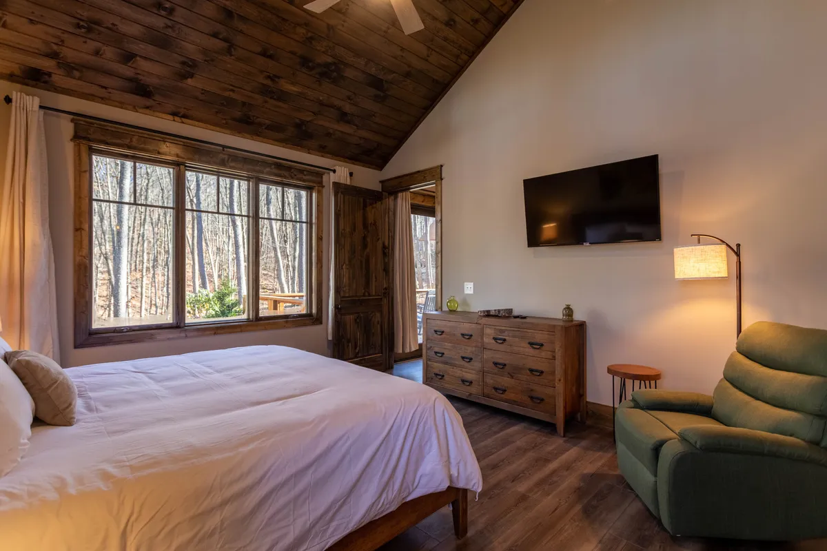 A serene master suite featuring a bed with crisp white bedding, a large window that frames calming views of nature, a sturdy wooden dresser, and a comfortable green chair for quiet moments. The space is styled to feel private and peaceful, perfect for unwinding after a day of adventure.