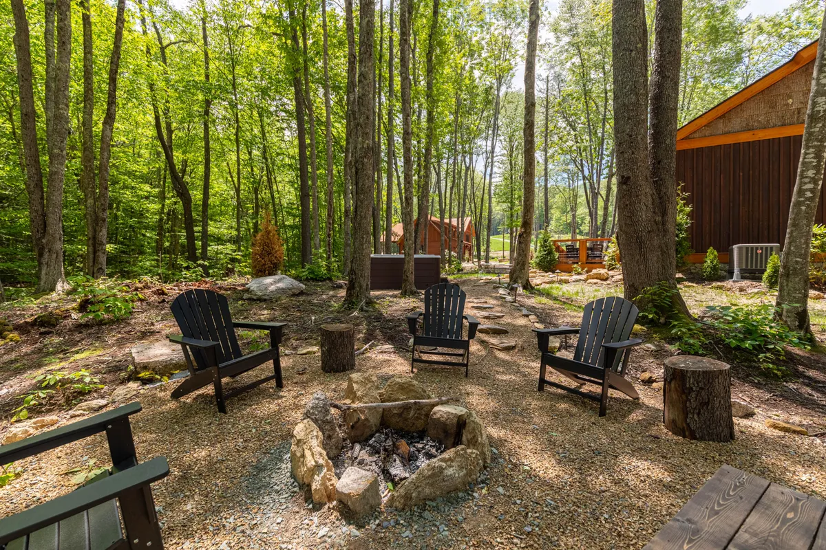 A cozy fire pit area with Adirondack chairs encircled by tall trees and a winding pathway. The arrangement invites gatherings for storytelling, roasting s’mores, or simply enjoying the starry nights. The woodland backdrop enhances the sense of peace and retreat.