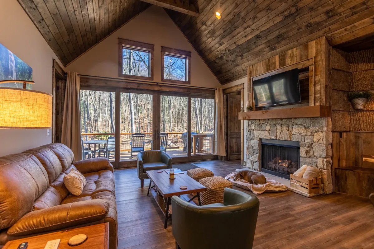 A cozy living room scene featuring large windows overlooking a forest view, a striking stone fireplace, plush leather sofas, and a wooden coffee table. The space feels both relaxing and social, perfect for enjoying coffee, unwinding by the fire, or gathering with friends.