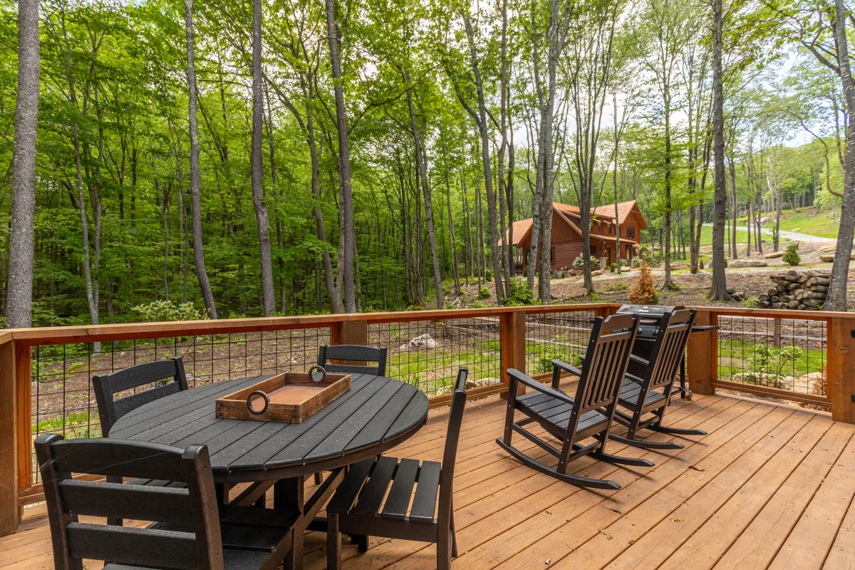 A spacious wooden deck equipped with rocking chairs, a circular dining table, and views of surrounding trees and the cabin in the background. The area is ideal for outdoor dining, relaxation, and social gatherings, seamlessly blending indoor comfort with natural beauty.
