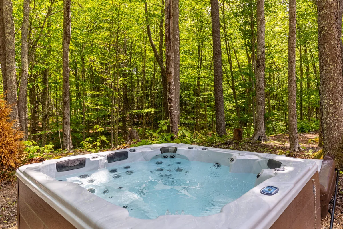 A serene exterior scene featuring a private hot tub nestled among lush, mature trees. The area is surrounded by natural foliage, creating a sense of privacy and immersion in nature. The tranquil setting is perfect for relaxing, stargazing, or enjoying a peaceful morning in the fresh mountain air.