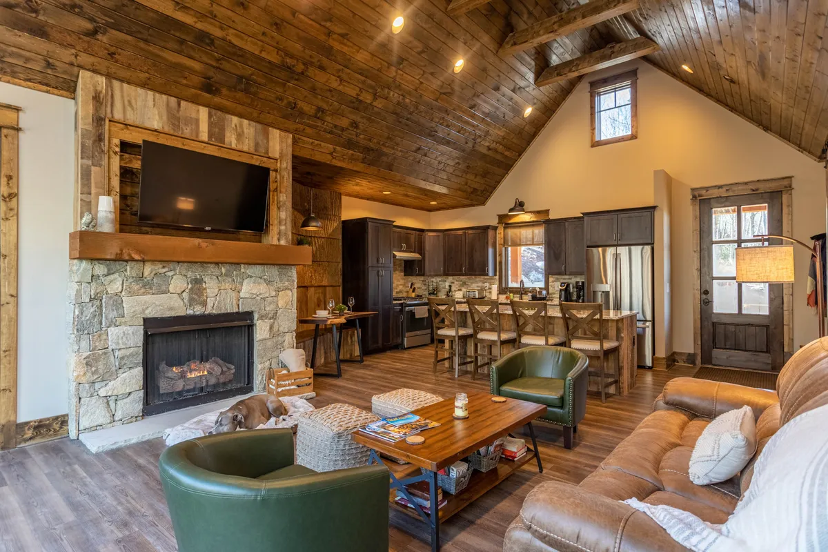 This image captures a warm and inviting open-concept living area featuring a prominent stone fireplace, an oversized TV, a spacious kitchen area, and comfortable seating. The scene suggests a space well-suited for gatherings, movie nights, and shared meals, with the layout fostering easy movement and conversation.