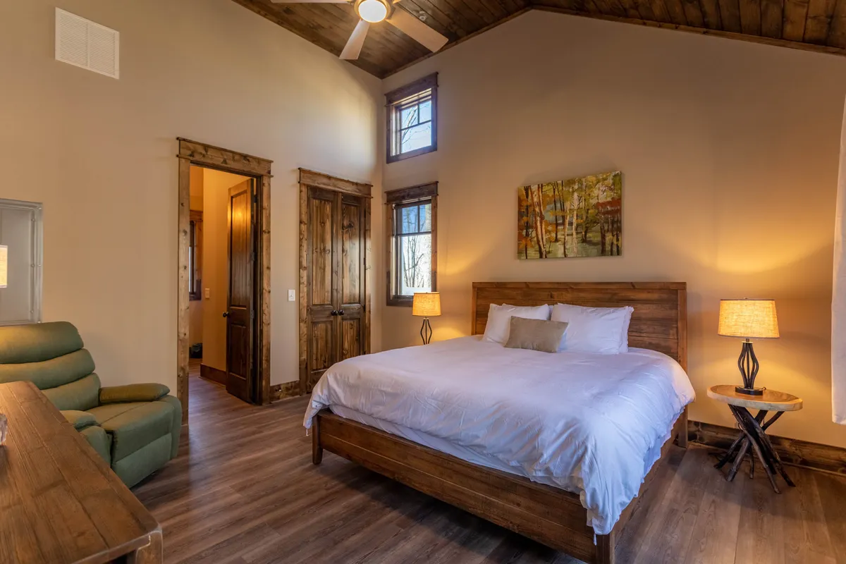A tranquil master bedroom featuring a king-sized bed adorned with crisp white linens, flanked by wooden nightstands and complemented by a cozy green armchair in the corner. Soft natural light filters in, creating an inviting atmosphere ideal for relaxation and restful sleep.